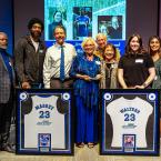 A group of nine people smiling for a photo at a jersey retirement ceremony, with two framed jerseys on display in the foreground.