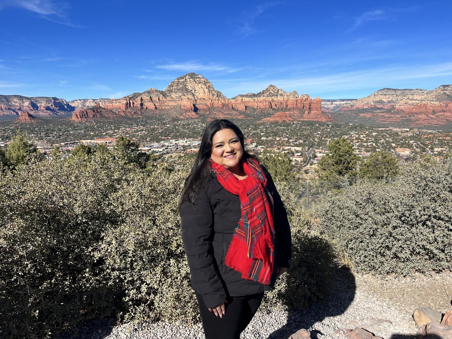 Edlin in a black coat and red scarf, in front of a wide canyon with a city below it.