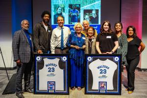 A group of nine people smiling for a photo at a jersey retirement ceremony, with two framed jerseys on display in the foreground.
