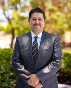 Dr. Roberto Gonzalez, the new president of Oxnard College, standing outdoors in a suit and tie, smiling with trees and greenery in the background.