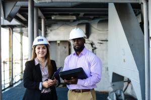 Two people wearing safety helmets, one holding a tablet and the other holding a clipboard, standing in an industrial setting.