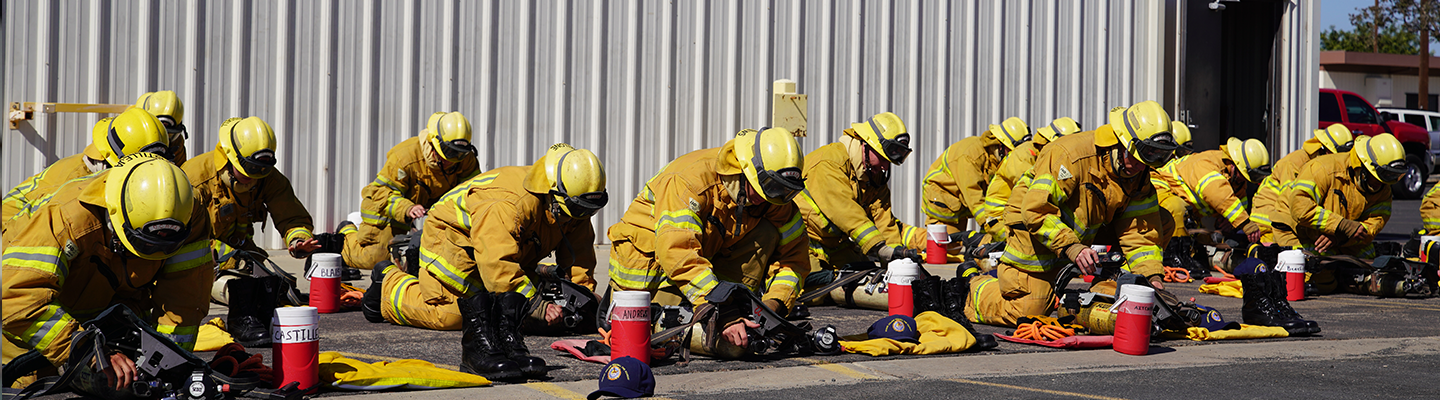 Fire academy students in full firefighter garb gathering tools from the ground.
