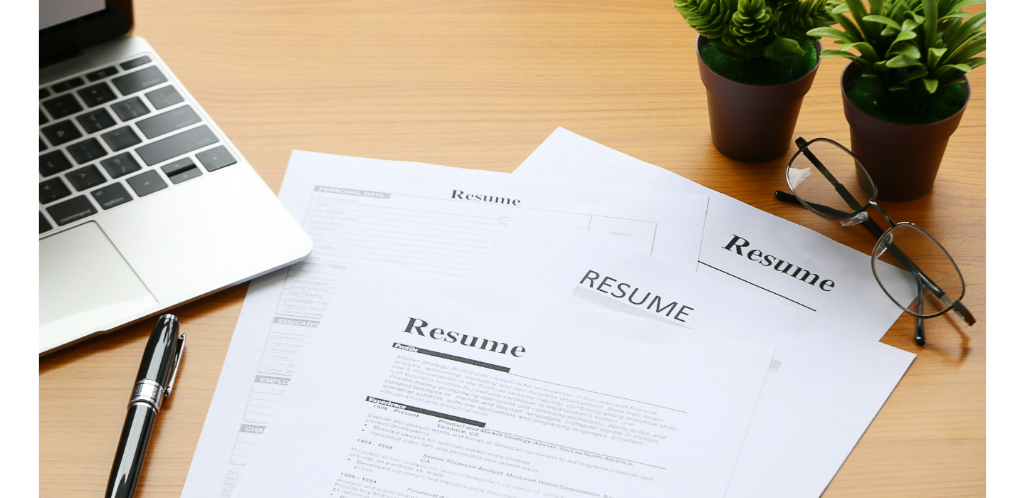 photo of tabletop with open laptop in upper left corner and black ballpoint pen on table just below laptop. In the center of the image are three printed pages with the word "Resume" at the top. Just above paperwork in upper right corner of image are two small indoor plants in terracotta pots with eye glasses sitting on table just below plants.