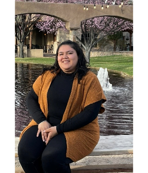 photo of Esmeralda Martinez-Rosales, Career Coach. Esmeralda is sitting on a stone bench in front of a water installation with a bridge and cherry blossom trees in the background.