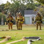 Four Fire Academy students standing outdoors in uniforms with a fire hose extended across the grass on the Oxnard College Public Safety Campus.
