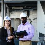 Two people wearing safety helmets, one holding a tablet and the other holding a clipboard, standing in an industrial setting.