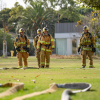 Four Fire Academy students standing outdoors in uniforms with a fire hose extended across the grass on the Oxnard College Public Safety Campus.