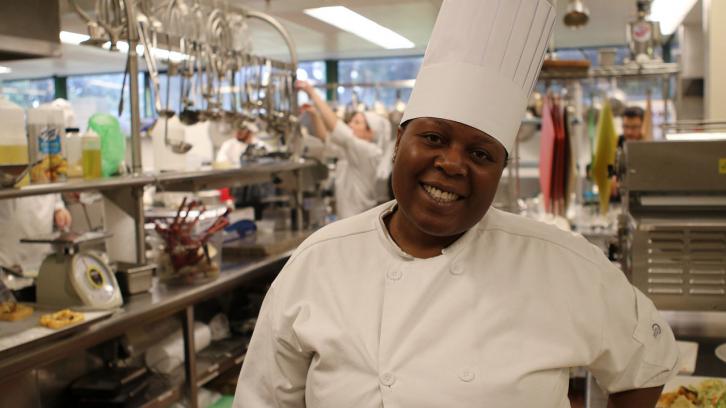 female Culinary student in uniform standing in foreground to Bistro kitchen.