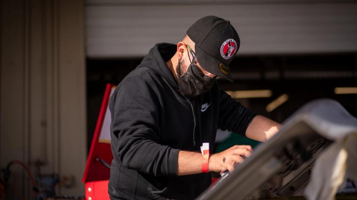 male student wearing COVID mask and baseball cap working on auto body project