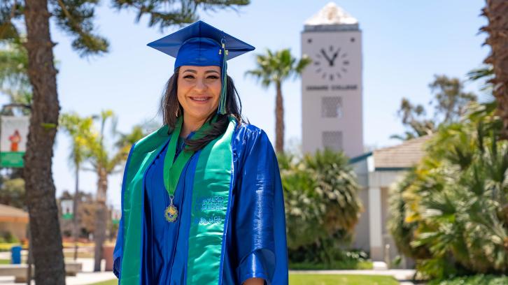 female student in blue graduation cap and gown, standing with Oxnard College clock tower in background