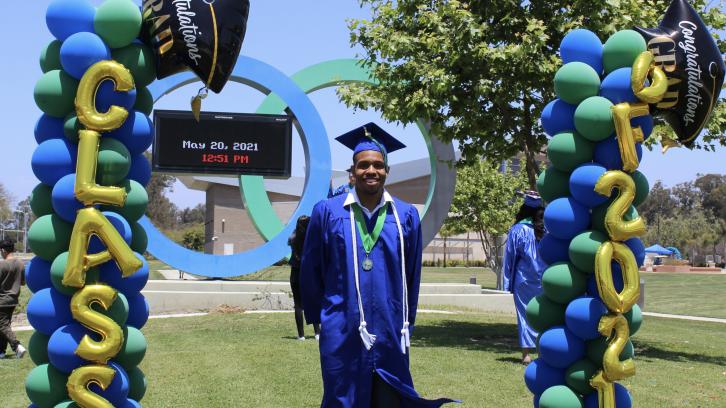 male student wearing blue graduation cap and gown, standing on campus lawn with "Class of 2021" balloons on either side, and OC marquee in background.