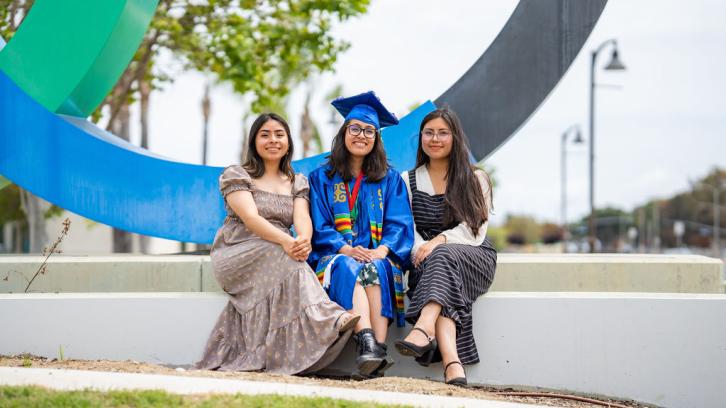 photo of Oxnard College graduate with female friends sitting on either side of student. All three individuals are in the foreground of the OC marquee sign.