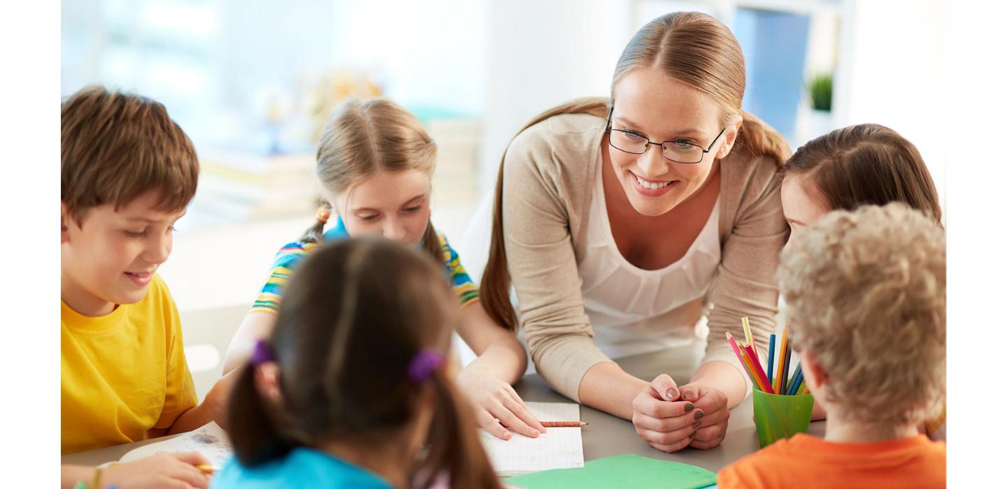 group of 5 young children sitting at table and interacting with female teacher