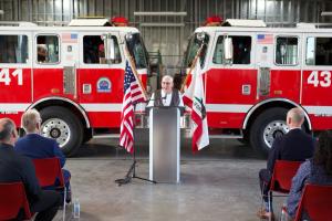 Oxnard College Fire Academy Founder Ed French speaks at dedication event standing at a podium in front of two fire engines, the American flag, and the California flag.